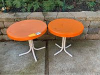 Two round orange metal patio tables side by side with white metal legs on concrete surface with stone wall background.
