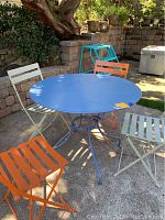 Blue round patio table with four foldable metal chairs in white and orange, set outdoors showing some wear.