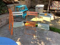 Four metal folding patio chairs (two orange, two light gray) displayed outdoors on a concrete surface with some lawn and stone wall background.