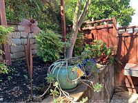 Photo showing turquoise potted succulent and metal flower yard art outdoors in garden area next to stone retaining wall and wooden fence.