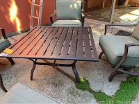 Square metal patio table with slatted top and four angled legs on outdoor concrete surface with moss patches visible.