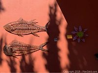 Three pieces of outdoor rusted metal wall art mounted on a reddish-brown wall: two fish sculptures each 31 inches long, and one flower sculpture approximately 12 inches in diameter.