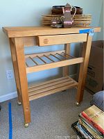 Front view of wooden kitchen cart with butcher block top, drawer, and lower slatted shelves. Casters visible.