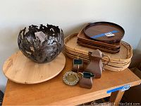 Seven bamboo trays, five wooden trays stacked, one circular Lazy Susan on the left, metal decorative bowl, metal creamer, sugar, and teapot set on wooden table surface.