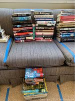 Photo showing three stacks of assorted books on a couch, including works by John Grisham, Tom Clancy, Stephen White, and others.