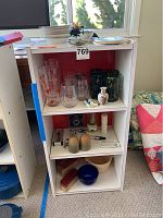 Three shelf white shelving unit displaying a variety of glassware and ceramics including fish glasses, art swirl glasses, plastic stemless wine glasses, ceramic salt and pepper shakers, wine opener, little Dutch clogs, and a small figurine.