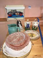 The entire lot arranged on a wooden surface showing pink pressed glass plates, floral ceramic plates, turquoise ceramic pitcher, and two small ceramic figurines of children with a ceramic dog statue behind them and a boxed Japanese decor in background.