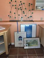 Metal wall art sculpture with multiple curved rods ending in green circular discs, standing on a kitchen counter with three framed artworks resting nearby.