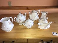 Seven porcelain pieces arranged on wooden table, showing floral tea pots, creamers, sugar bowls, and gravy boat.