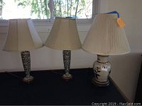 Three table lamps on a black surface with window background, showing shapes, designs, and shades, to document condition and style of lamps in lot.