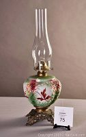 Full oil lamp showing floral decorated glass font with brass metal base and tall glass chimney.