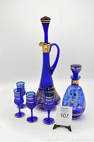 Full set of one tall blue decanter with stopper and handle, one smaller blue decorative decanter with stopper, and four matching blue stemmed glasses with gold accents.
