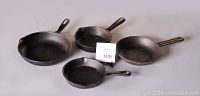 Four mixed vintage cast iron frying pans arranged side by side on a neutral surface, showing the cooking surfaces and integrated handles.