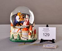 Snow globe with Beauty and the Beast figurines inside sitting on a castle base with multiple orange-roofed towers and green trees.
