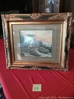 Full view of Thomas Kinkade print 'Seaside Retreat' in ornate gold carved frame on red cloth background with some damage visible on frame edges.