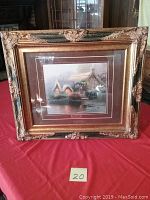 Framed artwork on a red cloth with ornate gold frame, showing a cottage scene with water and flowers.