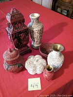 Ten oriental style decorative pieces including vases and bowls on red cloth background showing detailed carvings and varying colors red/black and cream/brown.