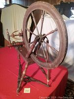 Front and side view of the antique wooden spinning wheel showing the large wheel, spindle, and base structure with aged wood and missing bobbin.