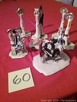 Group view of all 8 porcelain statues including bride and groom, seated couple, piano scene, and three single ladies with hats on red cloth surface.