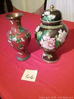 Two painted brass vases on red table, one red 12-inch and one black 13-inch with lid and floral paintings.