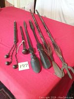 Six decorative weapons displayed on red cloth including two long spears with metal tips, two carved wooden spears with human heads, a spiked wooden club with chains, and a short wooden club with rounded end.