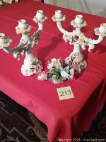 Pair of white porcelain candelabras with three candle holders each and floral decoration on a red tablecloth.