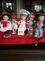 Five Campbell Kids dolls in a row on red cloth, various themes representing cooking, painting, beach, and others.