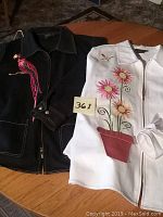 Two jackets on a wooden surface: white jacket with floral embroidery and black jacket with parrot embroidery.