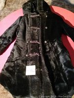 Front view of a small black faux fur coat with hood, frog-style closures, and snap buttons lying flat on a surface.