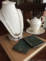 Full view of necklace on bust, two small antique books on a large book and the teacup and teapot set on saucer