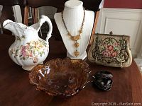 Full view of six vintage items on a wooden table: floral ceramic pitcher, amber and clear bead necklace on a bust, petit point purse, amber glass dish, and two black floral bobbie pins.