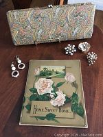 Front view of vintage paisley clutch showing multicolor pattern, three brooches and artisan earrings arranged beside Home Sweet Home print.
