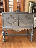 Front view of the vintage cabinet showing ornate carved details, double doors with brass knobs, and turned legs.