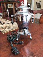 Full view of vintage electric percolator coffee pot on wooden table, showing polished metal body, black handles, power cord, and legs.