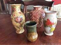 Four decorative vases arranged on table showing cranberry and gold floral vase, tan floral vase behind greenery colored pottery vase and vase with autumn leaves