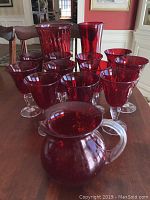 Eleven ruby/cranberry stemware glasses with clear stems and one small pitcher with a clear handle on a wooden table.