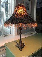 Lamp lit showing warm glow through black lace floral pattern on lampshade with fringed and beaded trim.