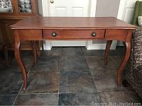 Front view of wooden desk showing the cabriole legs, single drawer with two round knobs, and rectangular top with decorative edges.