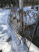 Three 5-foot rusty metal garden fence sections leaning against a vertical post outdoors in snow, showing curled top arches and lattice bottom with fleur de lis finials.