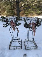 Side-by-side view of two metal pedestal plant stands in snow, decorated with winter greenery and ribbons.