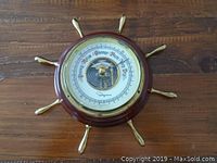 Full view of the round nautical style barometer with wooden frame and brass handles resembling a ship's wheel, showing the dial and external design.