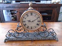 Full view of the entire metal table clock showing the antique-style face, bezel, and decorative scrollwork metal base.