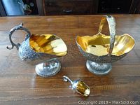 Front view of silver plated creamer, sugar basket with handle, and scoop on wooden table showing gold-tone interiors and embossed floral designs