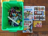 Green plastic bin with assorted Lego pieces and four Lego-themed books around it on the floor