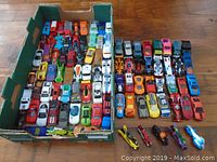 Top-down view showing over 90 die cast cars, trucks, and motorcycles arranged in multiple rows within a green cardboard box