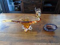 Wide view of the large amber-colored glass bird and small glass dish on a wooden surface in front of a wooden cabinet.