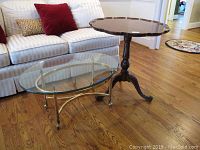 Two tables, glass coffee table with brass base in front and wood drop-leaf table behind on hardwood floor, white sofa and red pillows in background.