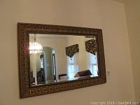 Full frontal view of rectangular wall mirror with ornate gold frame showing reflection of interior curtains and archways.