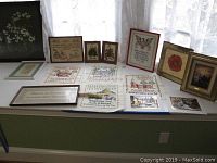 Several embroidered pieces and framed prints arranged on a window ledge, showing various embroidery designs and texts.