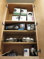 Light bulbs stored in wooden cabinet: boxes of LED bulbs, fluorescent bulb packaging, loose bulbs on middle shelf.